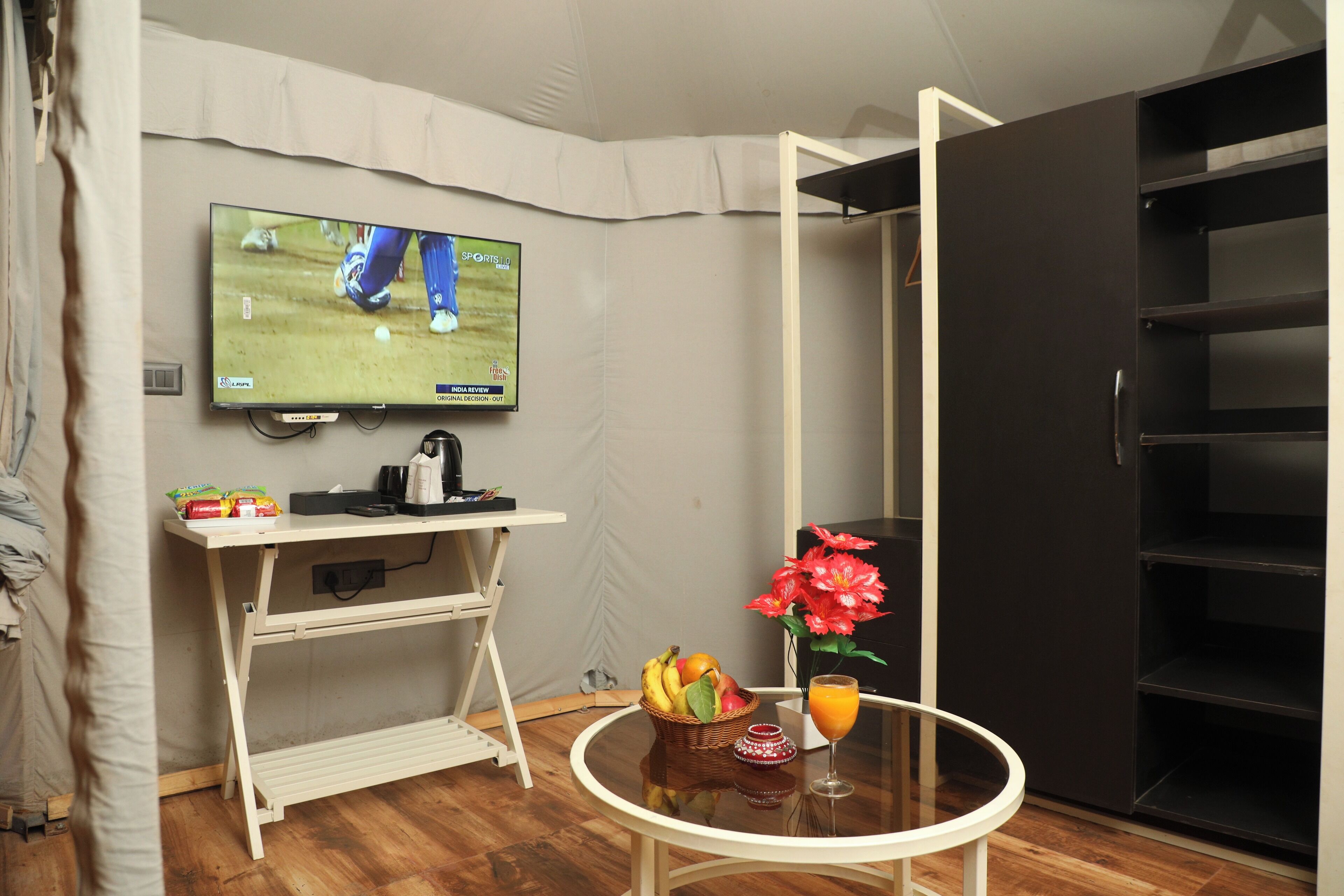Een hotelkamer met een televisie aan de muur, een kleine tafel met fruit en een drankje, een wit bureau en een zwarte garderobe.