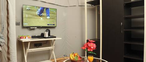 Een hotelkamer met een televisie aan de muur, een kleine tafel met fruit en een drankje, een wit bureau en een zwarte garderobe.
