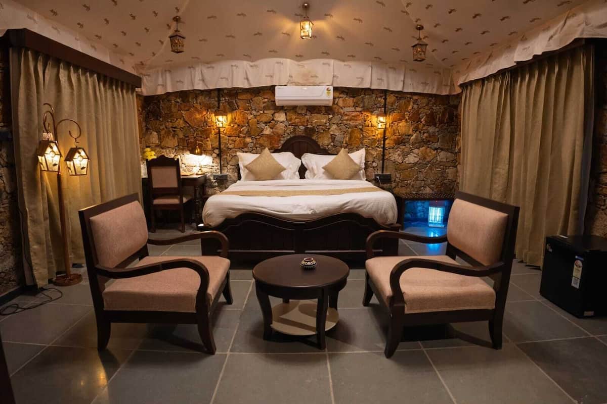 A hotel room with a stone wall, a large bed with multiple pillows, two armchairs, a small table, and a heater.