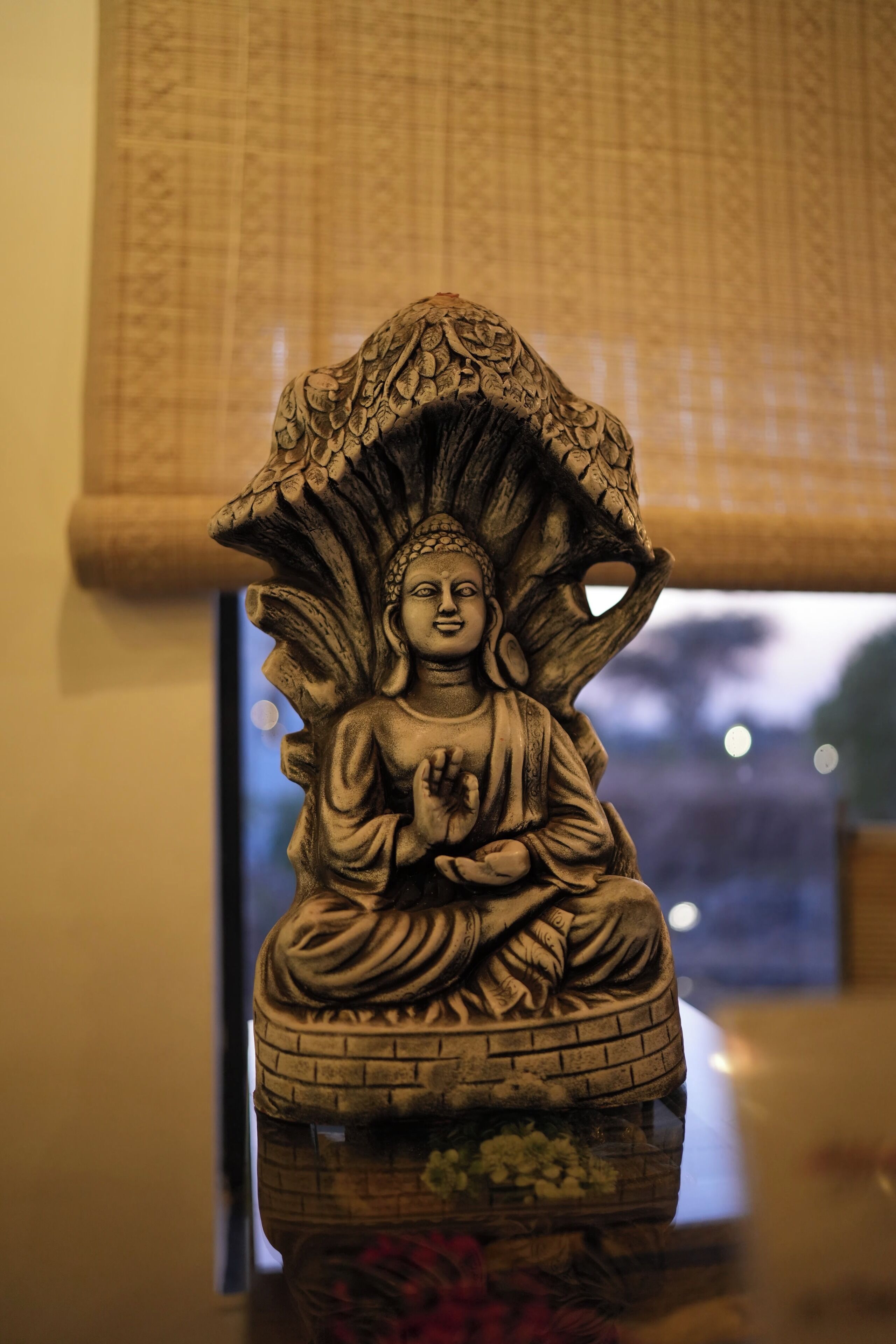 Una estatua de una figura sentada, con una base floral, ubicada en un espacio interior con un papel tapiz y una ventana al fondo.