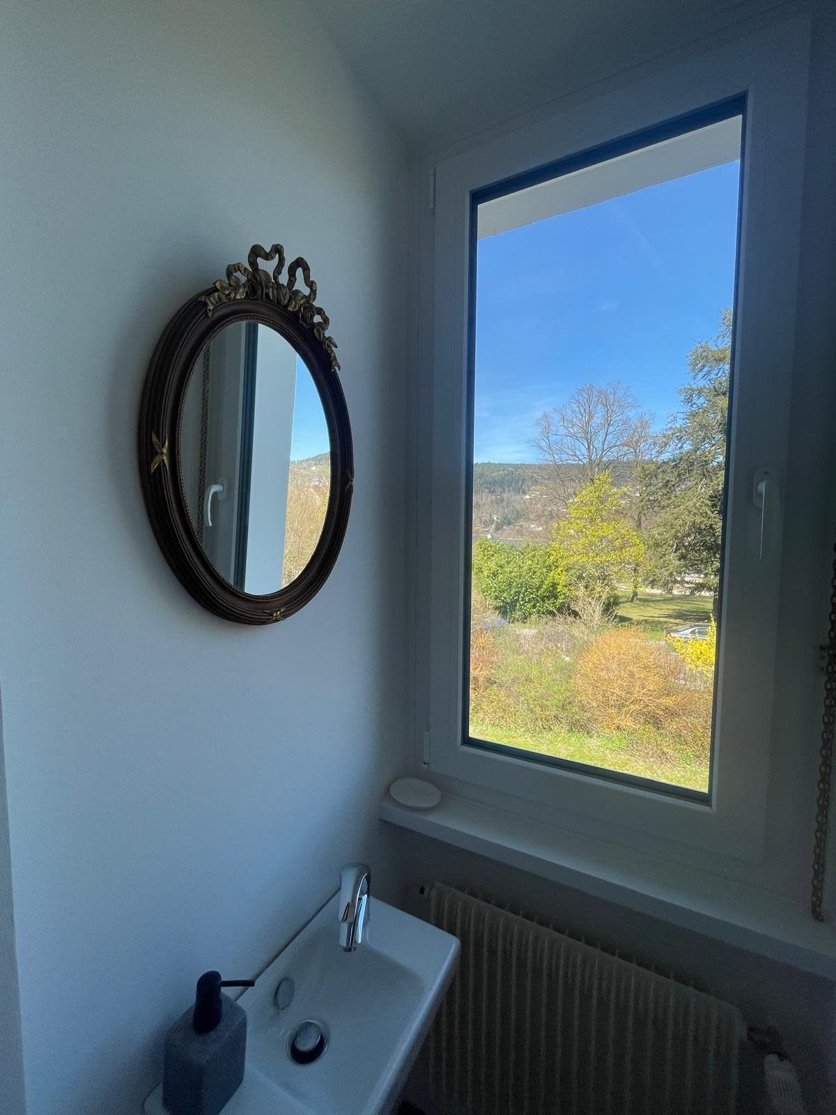 Un baño con un amplio ventanal, un espejo redondo y un lavamanos blanco.