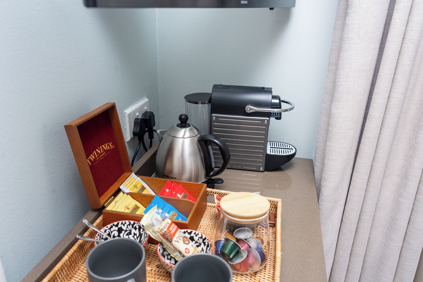 Una kitchenette con cafetera, hervidor y tetera. Hay una bandeja con tazas, una caja de bolsitas de té y un cesto con utensilios.