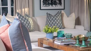 Una sala de estar acogedora con un sofá, almohadones estampados, una mesa de centro con libros y adornos, y una ventana con cortinas.