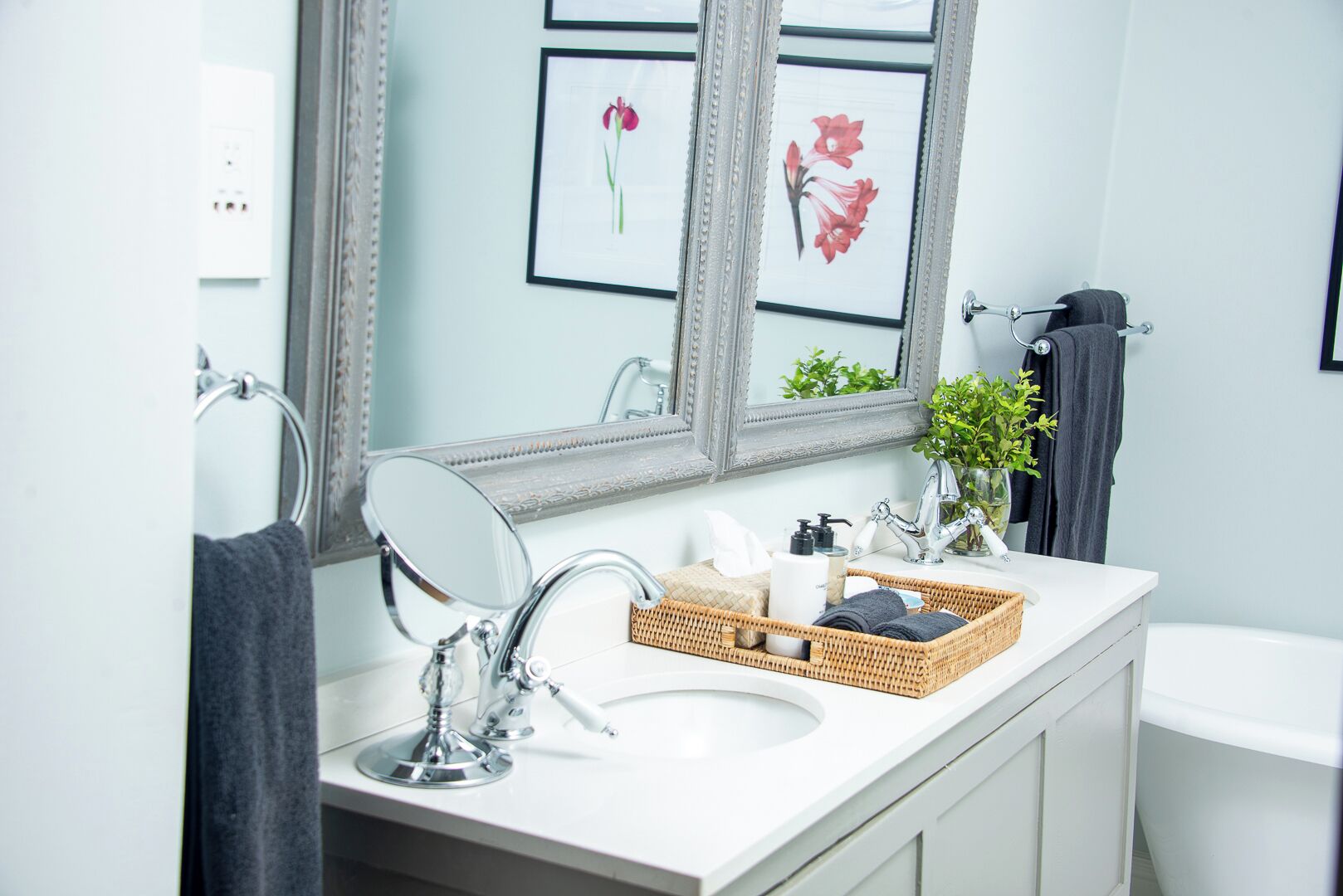 Une salle de bain moderne avec un lavabo blanc, un grand miroir et des œuvres d’art florales encadrées accrochées au mur.