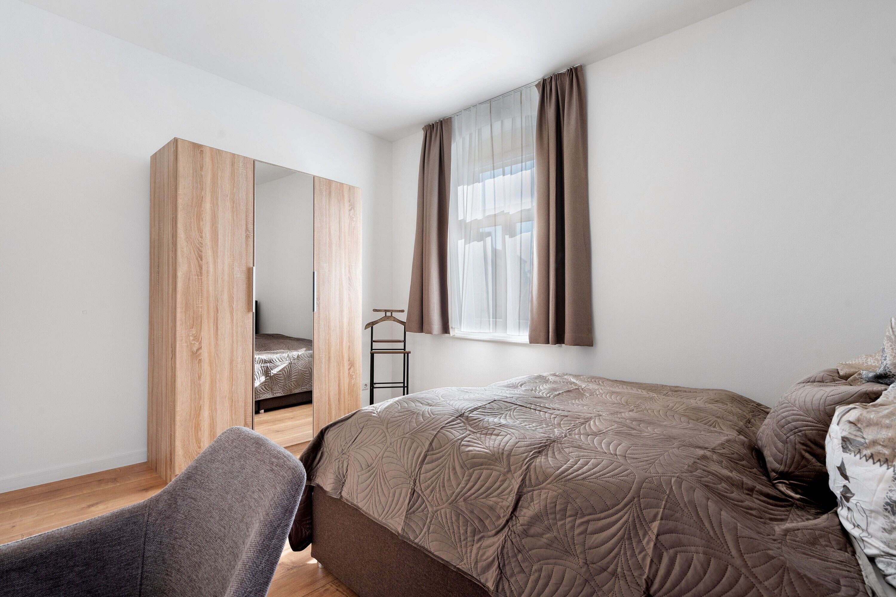 A bedroom with a bed, a chair, a wardrobe, and a window with curtains.