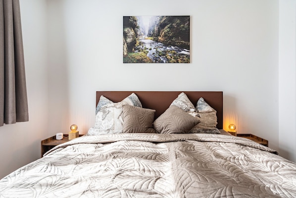 Une chambre à coucher avec un lit, des oreillers, une table de chevet avec une lampe et un tableau représentant une cascade, accroché au mur.