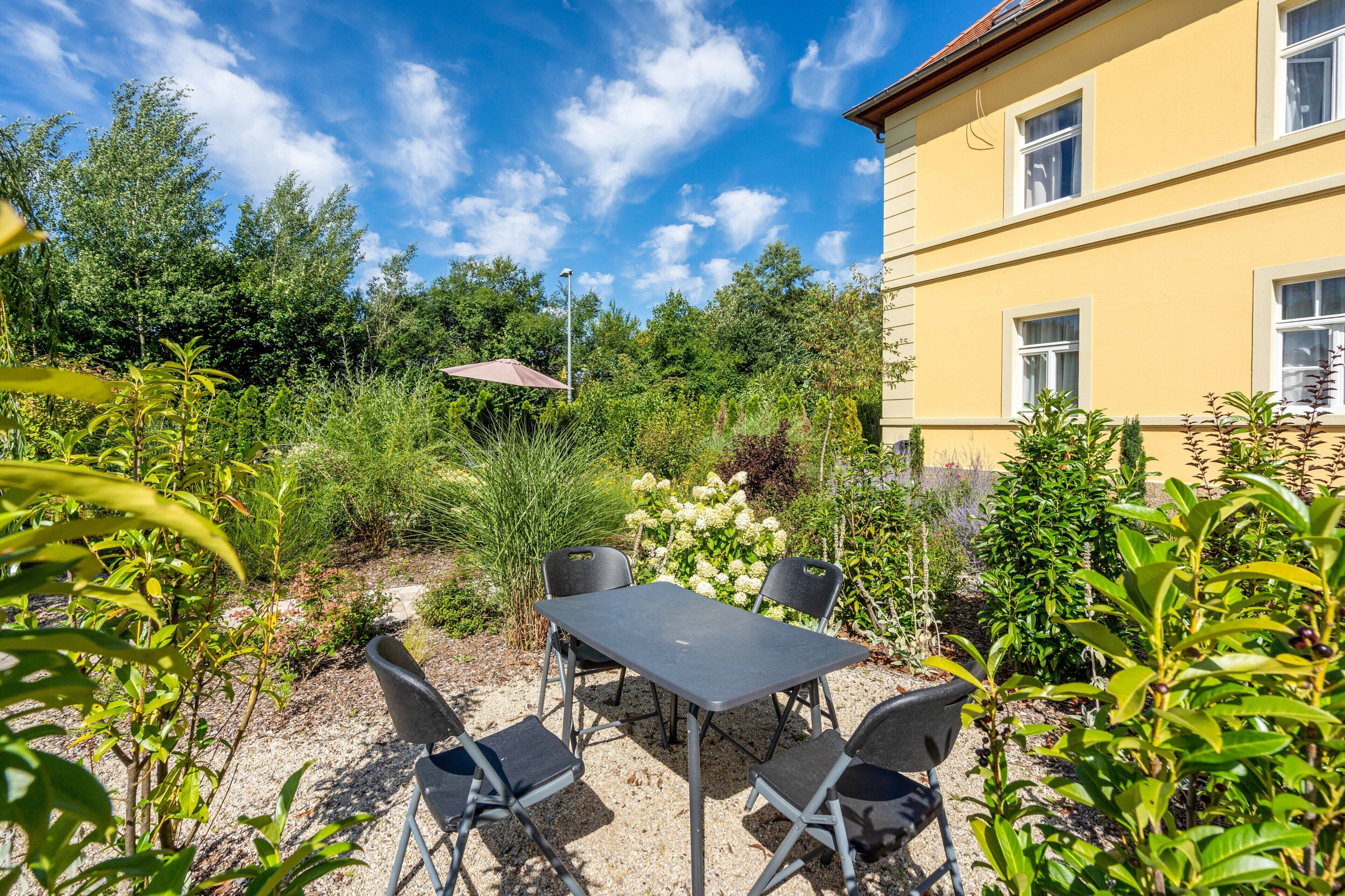 Ein Sitzbereich im Freien mit Tisch und Stühlen, umgeben von üppigem Grün und einem Gebäude im Hintergrund.