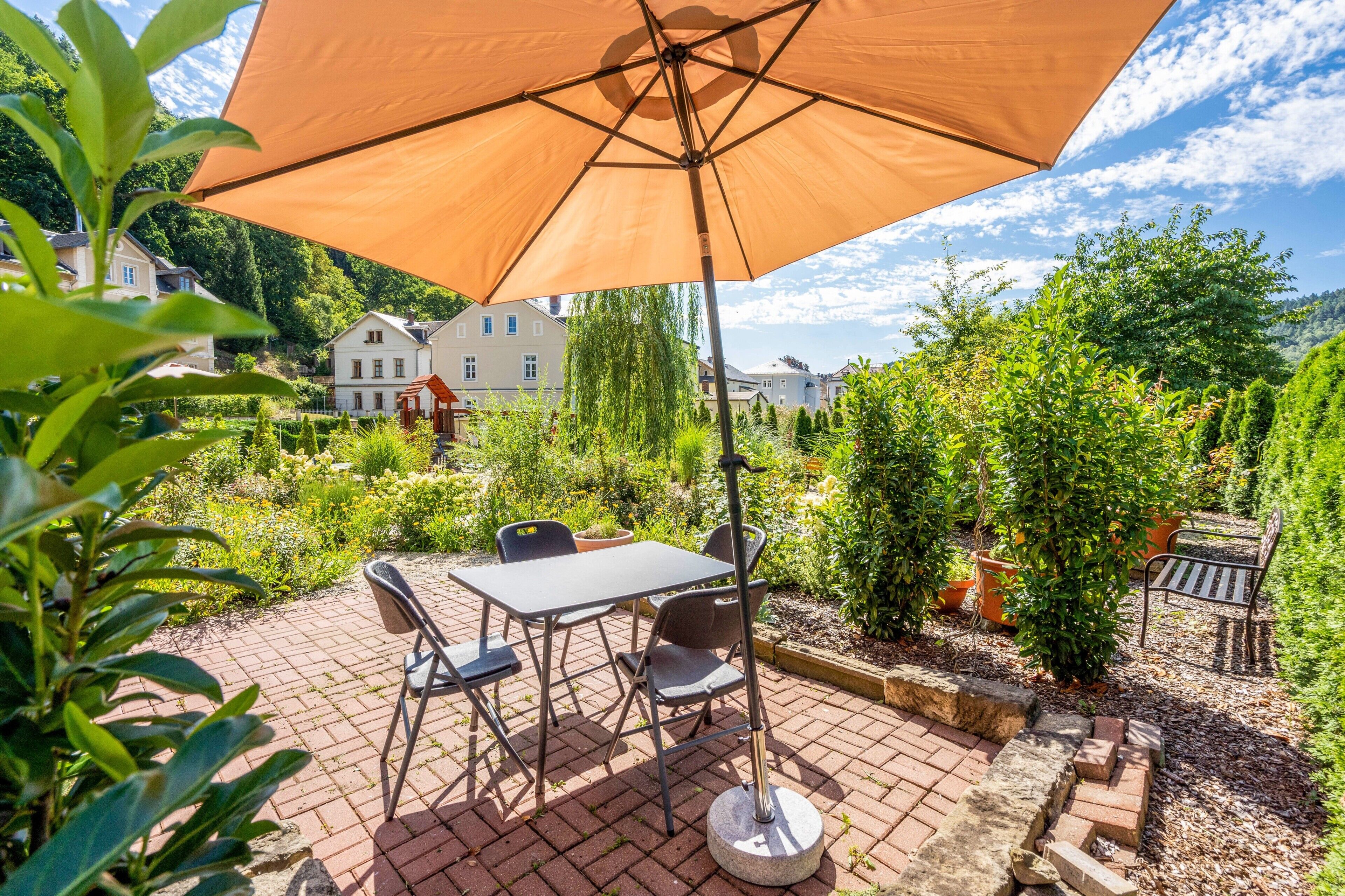 An outdoor patio with a table and chairs, surrounded by greenery and a view of houses in the background.