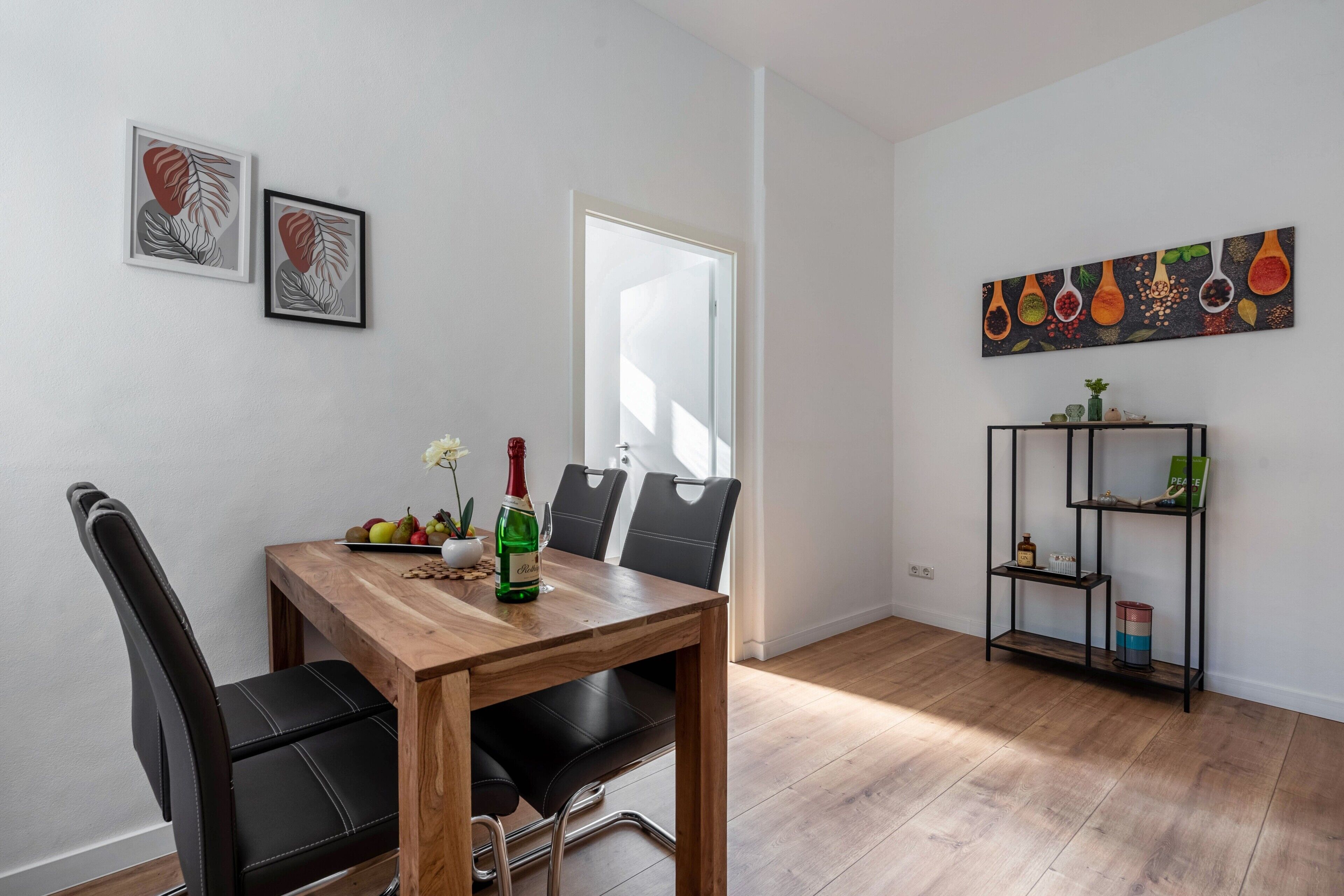 Ein Essbereich mit Holztisch, schwarzen Stühlen, einer Weinflasche und einer Obstschale. An der Wand hängen zwei gerahmte Kunstwerke, ein Bücherregal und eine Tür.