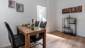 A dining area with a wooden table, black chairs, a bottle of wine, and a fruit bowl. There are two framed artworks on the wall, a bookshelf, and a door.