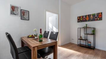 Ein Essbereich mit Holztisch, schwarzen Stühlen, einer Weinflasche und einer Obstschale. An der Wand hängen zwei gerahmte Kunstwerke, ein Bücherregal und eine Tür.