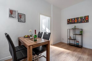 A dining area with a wooden table, black chairs, a bottle of wine, and a fruit bowl. There are two framed artworks on the wall, a bookshelf, and a door.