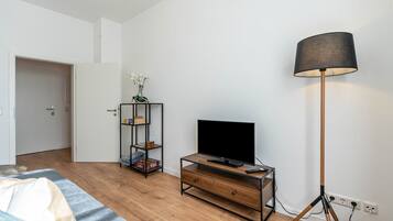 Una camera da letto con pavimento in legno, un letto con copriletto bianco e blu, una TV su un mobile in legno, una lampada da terra e una libreria con una pianta.
