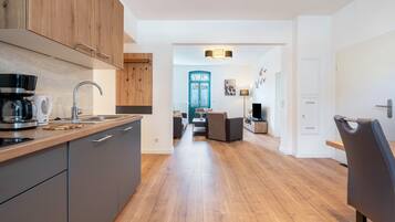 A modern kitchen with wooden cabinets, a coffee maker, and a sink. There is a dining area with a sofa and a table. The room has a wooden floor and white walls.