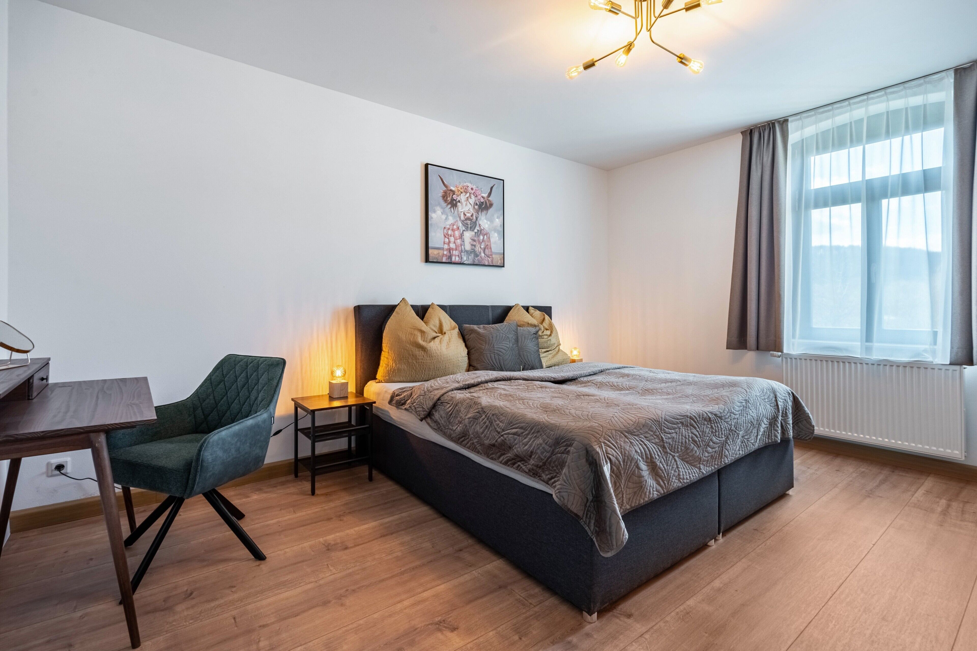 Une chambre à coucher comprenant un lit, un bureau, une chaise, une table de chevet, une lampe, un tableau accroché au mur et une fenêtre avec des rideaux.