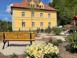 A yellow building with multiple windows, a bench, and a garden area with flowers.