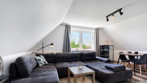Un salon moderne avec un canapé d’angle, une petite table, un coin repas avec des chaises et une grande fenêtre avec des rideaux.