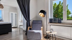 A modern room with a bed, a chair, a standing lamp, a small table, and a window with a view of trees. - Apartment 'Links' with Private Terrace and Private Garden (Wesselburenerkoog)