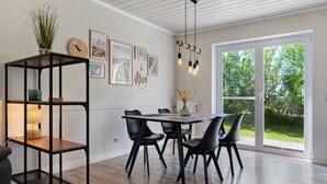 A dining area with a wooden table, black chairs, a black metal shelf, and a window with a view of greenery. - Apartment 'Links' with Private Terrace and Private Garden (Wesselburenerkoog)