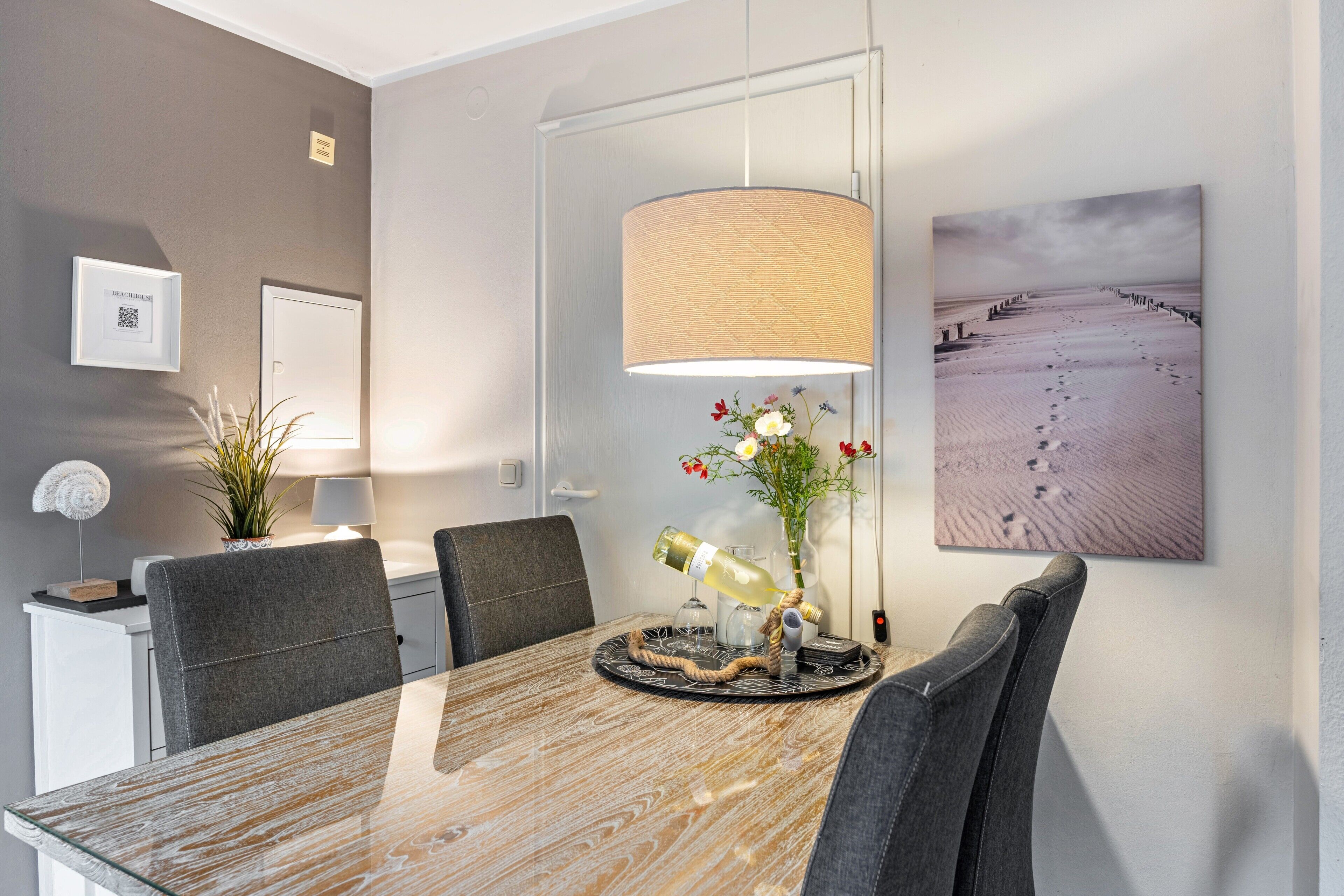 A modern dining area with a marble table, grey upholstered chairs, a hanging lamp, a framed artwork, and a small potted plant.