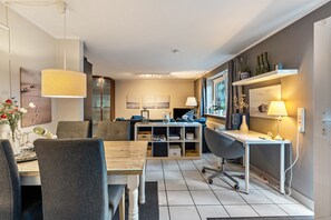 A modern living room with a dining table, chairs, a sofa, a desk, and a computer. There is a bookshelf, a window with curtains, and a hanging light fixture.