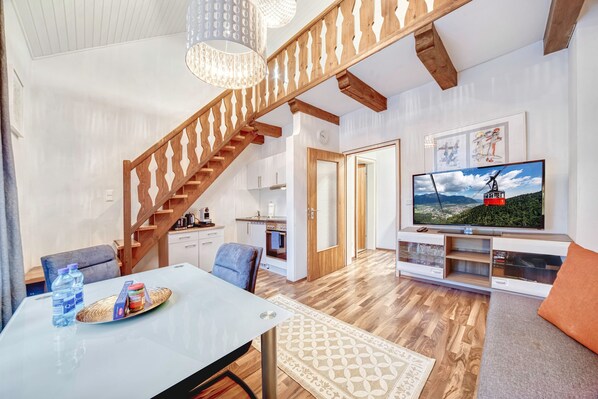 A modern living room with a wooden staircase, a dining area with a table and chairs, a kitchen area, and a TV mounted on the wall.