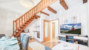 A modern living room with a wooden staircase, a dining area with a table set for four, a kitchen in the background, and a large TV displaying a mountain landscape.