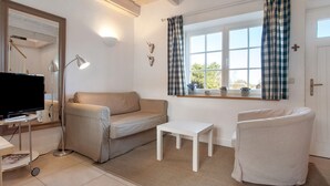 A living room with a sofa, a small table, a TV, and a window with curtains. - Holiday Apartment 'Jägerkate' with Private Terrace, Shared Garden and Wi-Fi (Walkendorf)