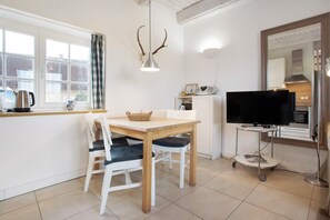 Un coin repas avec une table, des chaises, un téléviseur posé sur un meuble, une fenêtre avec des rideaux et, au fond, une cuisine visible.