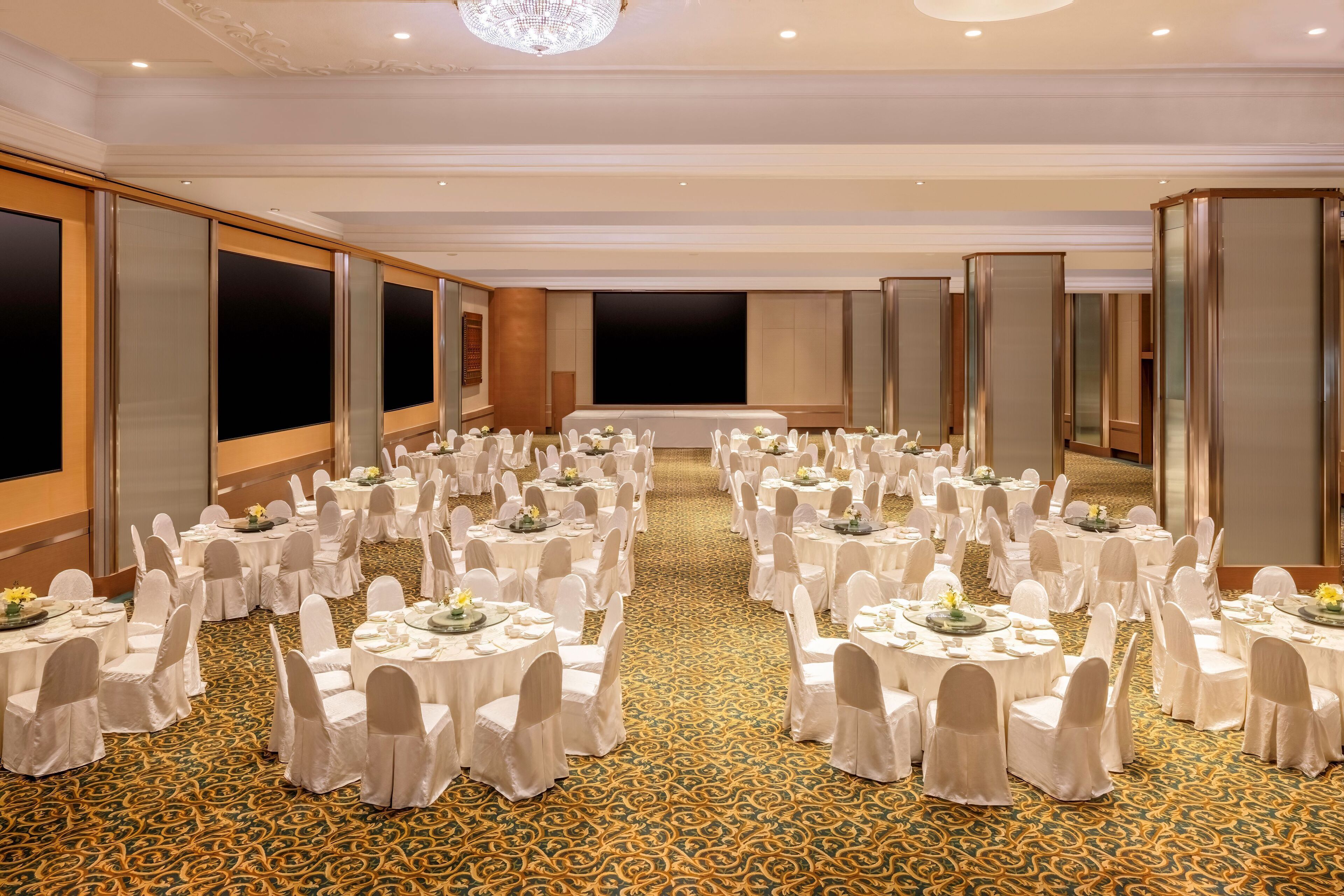 Un salón de banquetes con mesas redondas, sillas blancas y una alfombra estampada.