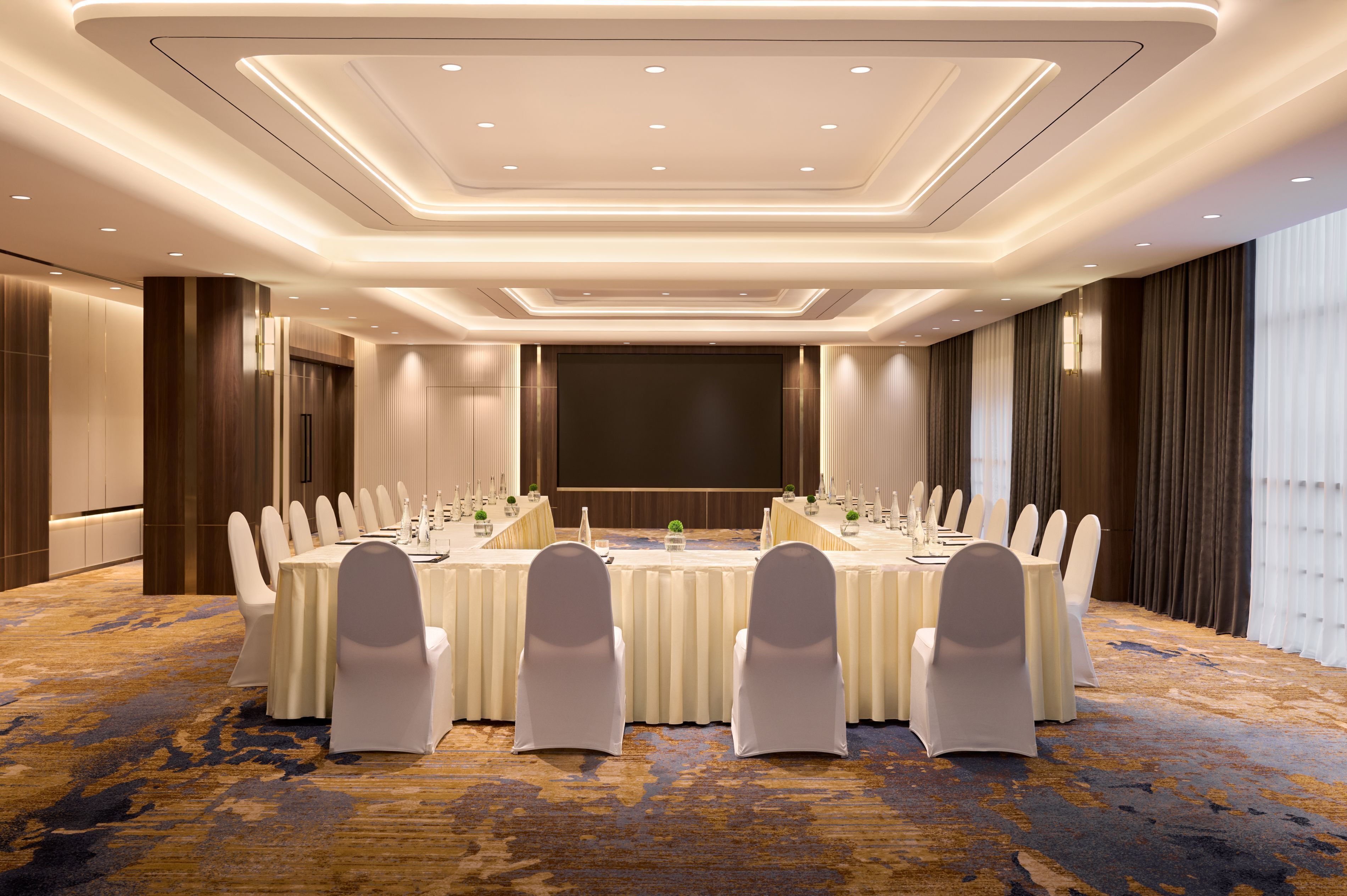 Un salón de conferencias con una larga mesa, sillas blancas y una pantalla grande.
