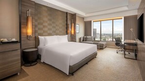 Ein modernes Hotelzimmer mit einem großen Bett, einem Schreibtisch, einem Stuhl und einem Fernseher. Das Zimmer bietet einen Blick auf die Stadt durch große Fenster.