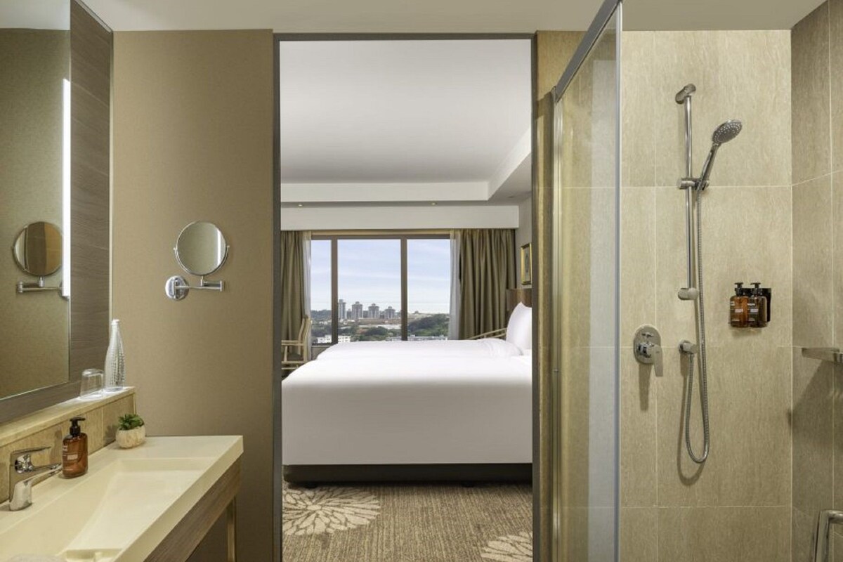 A hotel room with a large bed, a bathroom with a shower, a sink, and a view of the city.