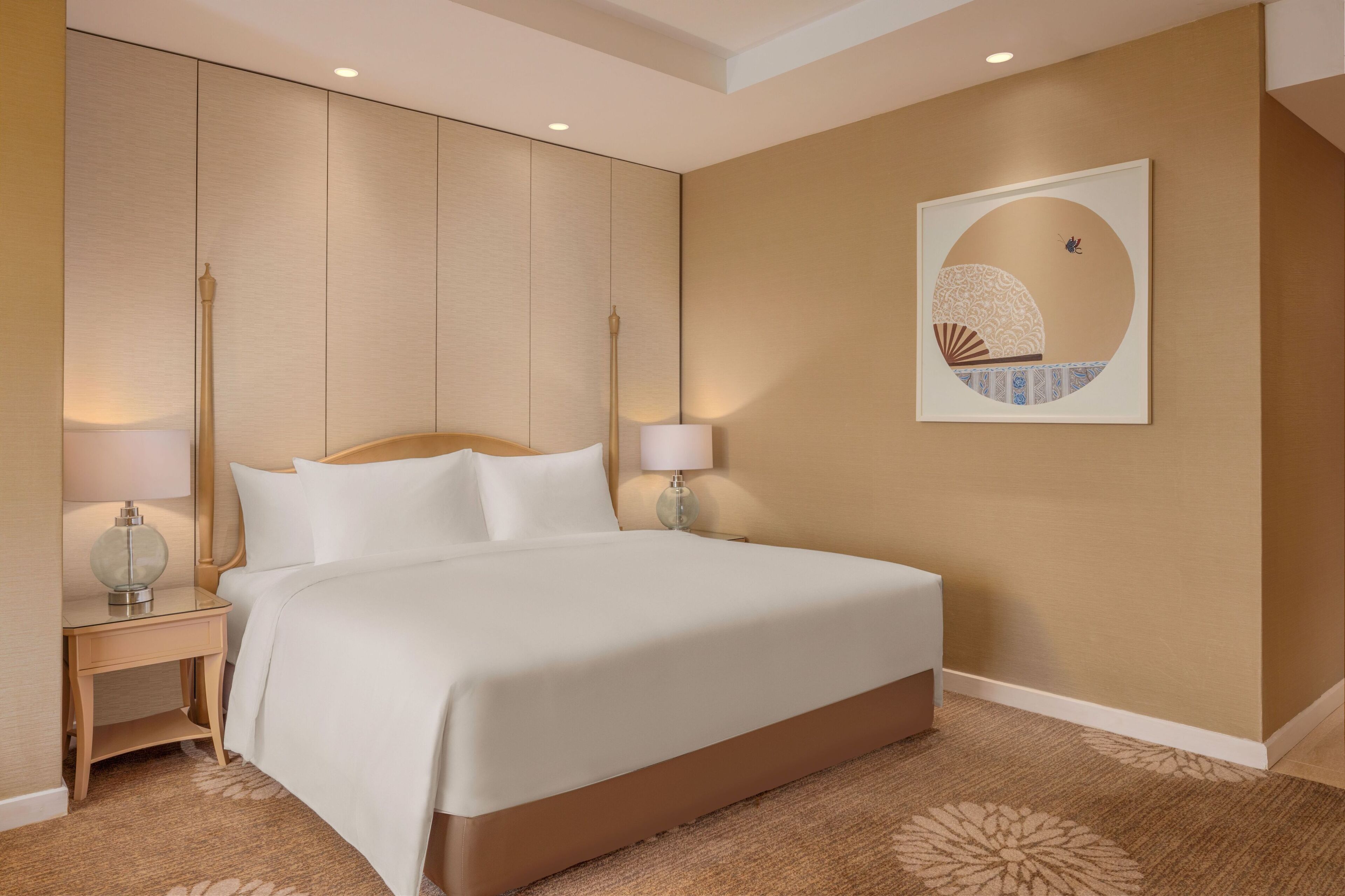 Habitación de hotel con una cama grande, dos mesitas de noche con lámparas, una obra de arte en la pared y piso alfombrado.