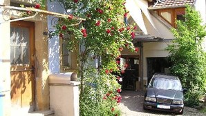 Un bâtiment devant lequel est garée une voiture, avec une porte et un petit jardin agrémenté de fleurs rouges.
