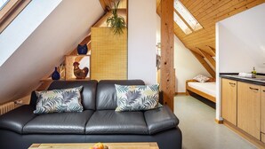 Un appartement moderne de style « loft », doté d’un canapé en cuir noir, d’une table à manger en bois agrémentée de fruits, d’un coin cuisine et d’un plafond en pente avec des poutres en bois.
