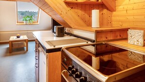 Une cuisine aux murs en bois, avec un poêle, une fenêtre ornée d’une plante et une petite table sur laquelle se trouve un bol.
