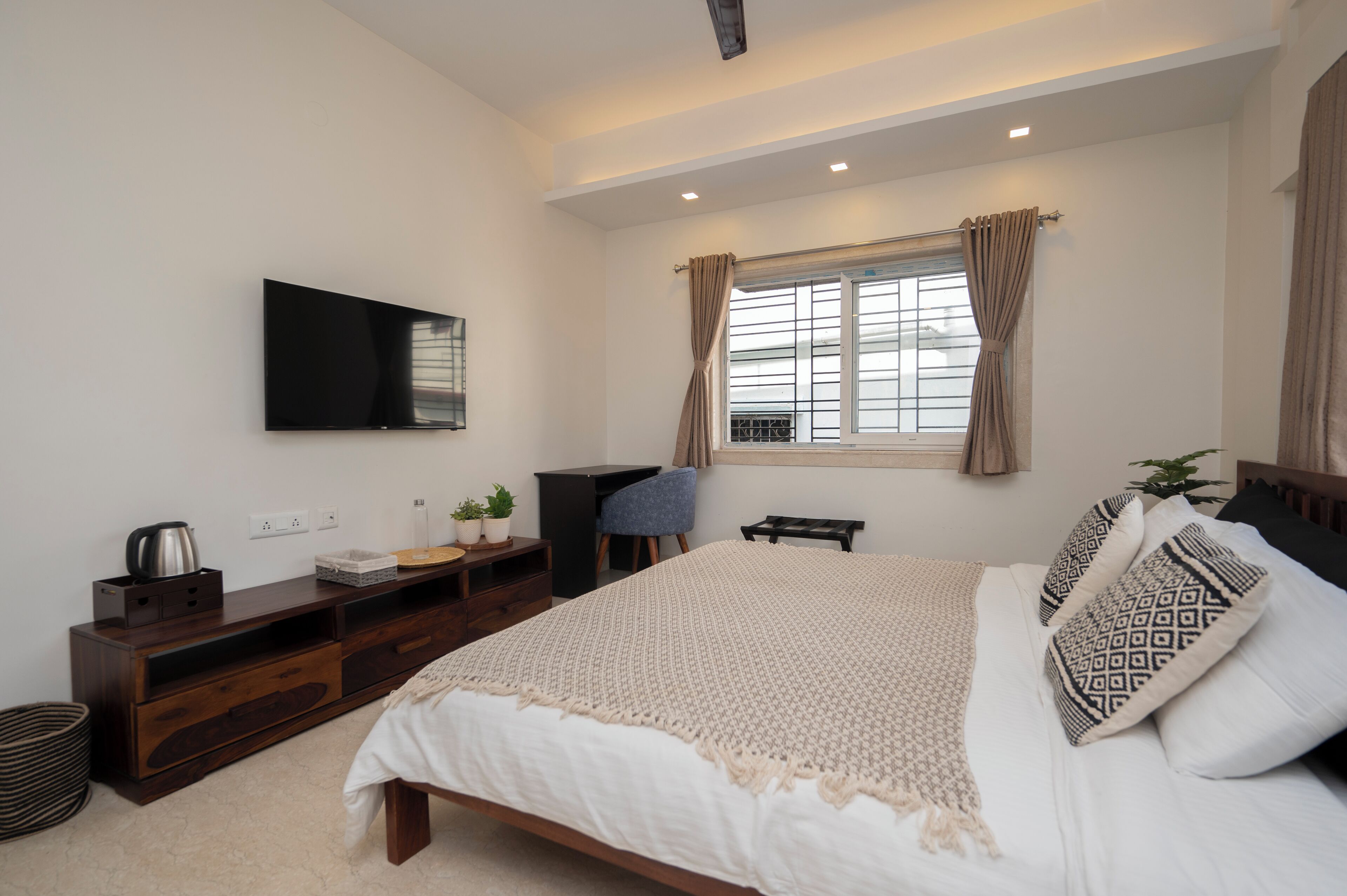 A bedroom with a bed, a TV mounted on the wall, a desk, and a window with curtains.