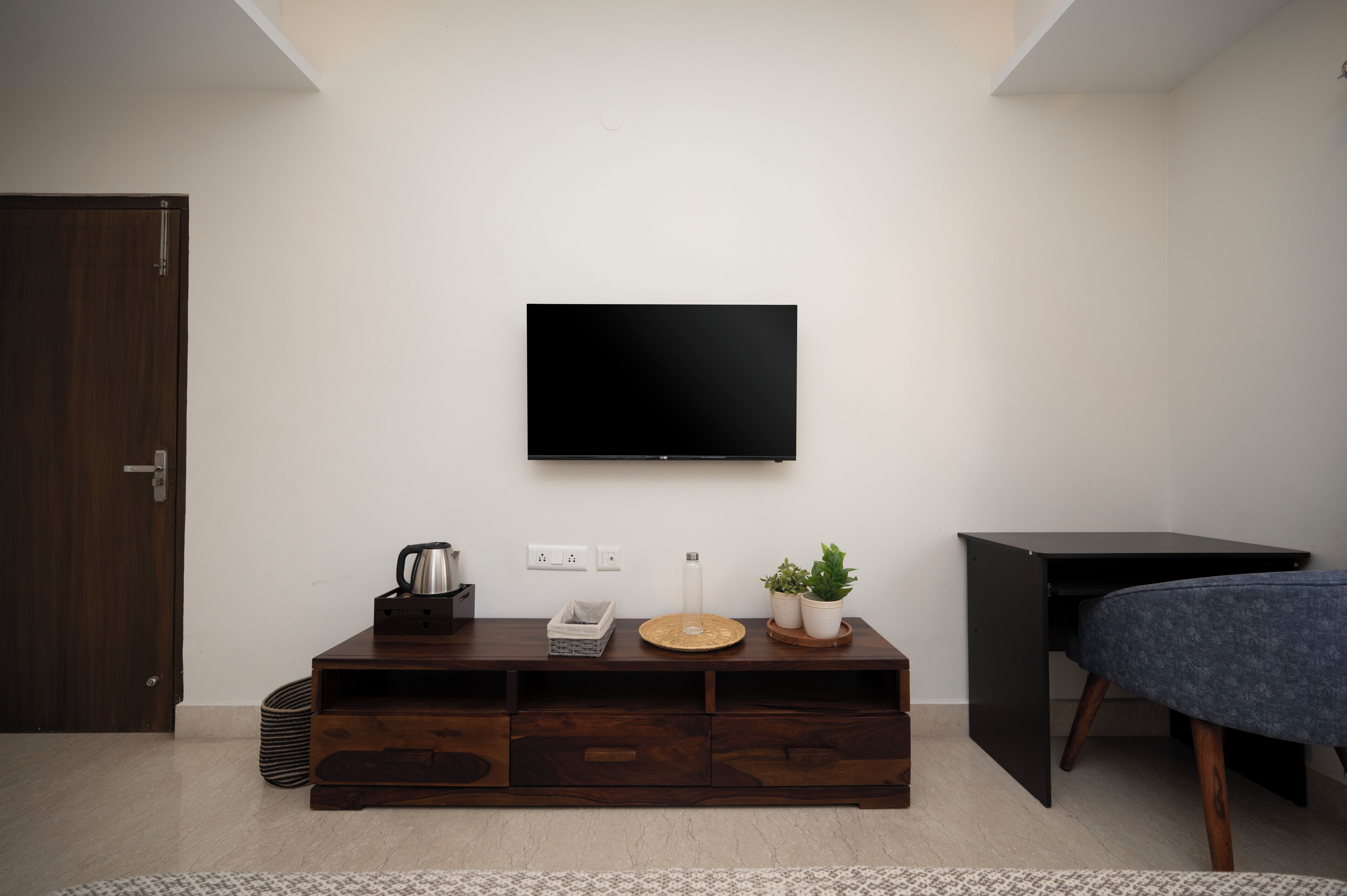 Quarto moderno com mesa de consola de madeira, televisão de ecrã plano na parede, uma pequena planta em vaso e uma cadeira estofada azul.