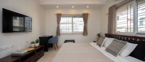 A bedroom with a bed, a desk, a chair, a TV, and a window with curtains.