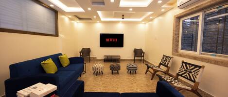 A living room with a blue sofa, two armchairs, a coffee table, and a TV mounted on the wall displaying 'NETFLIX'.