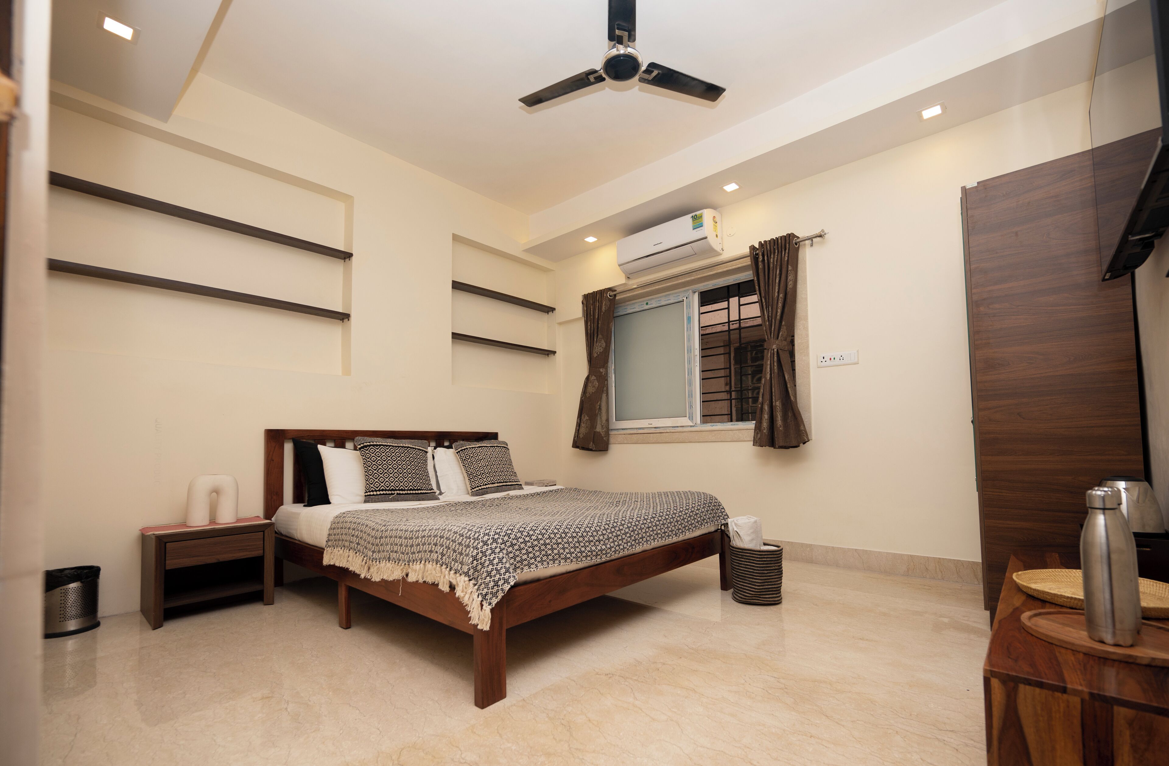 Un dormitorio con cama, mesitas de noche, ventilador de techo, equipo de aire acondicionado, ventana con cortinas y armario de madera.