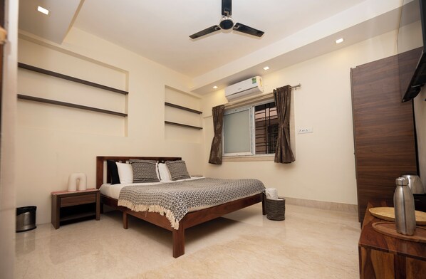 A bedroom with a bed, bedside tables, a ceiling fan, an air conditioning unit, a window with curtains, and a wooden wardrobe. - LAKESIDE SUITES (Kolkata)