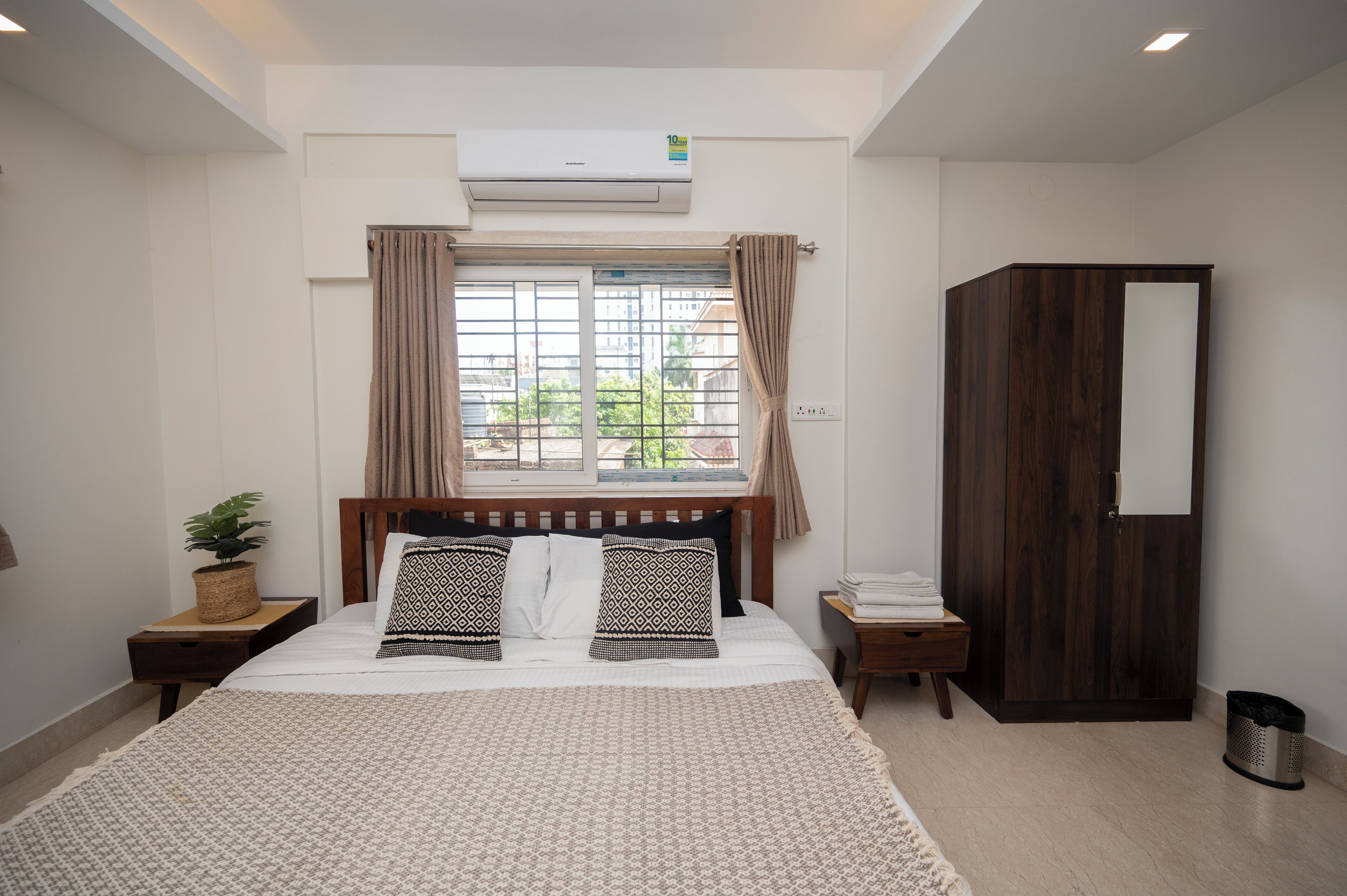 Un dormitorio con cama, ventana con cortinas, equipo de aire acondicionado, armario, mesita de noche con planta y cubo de basura.