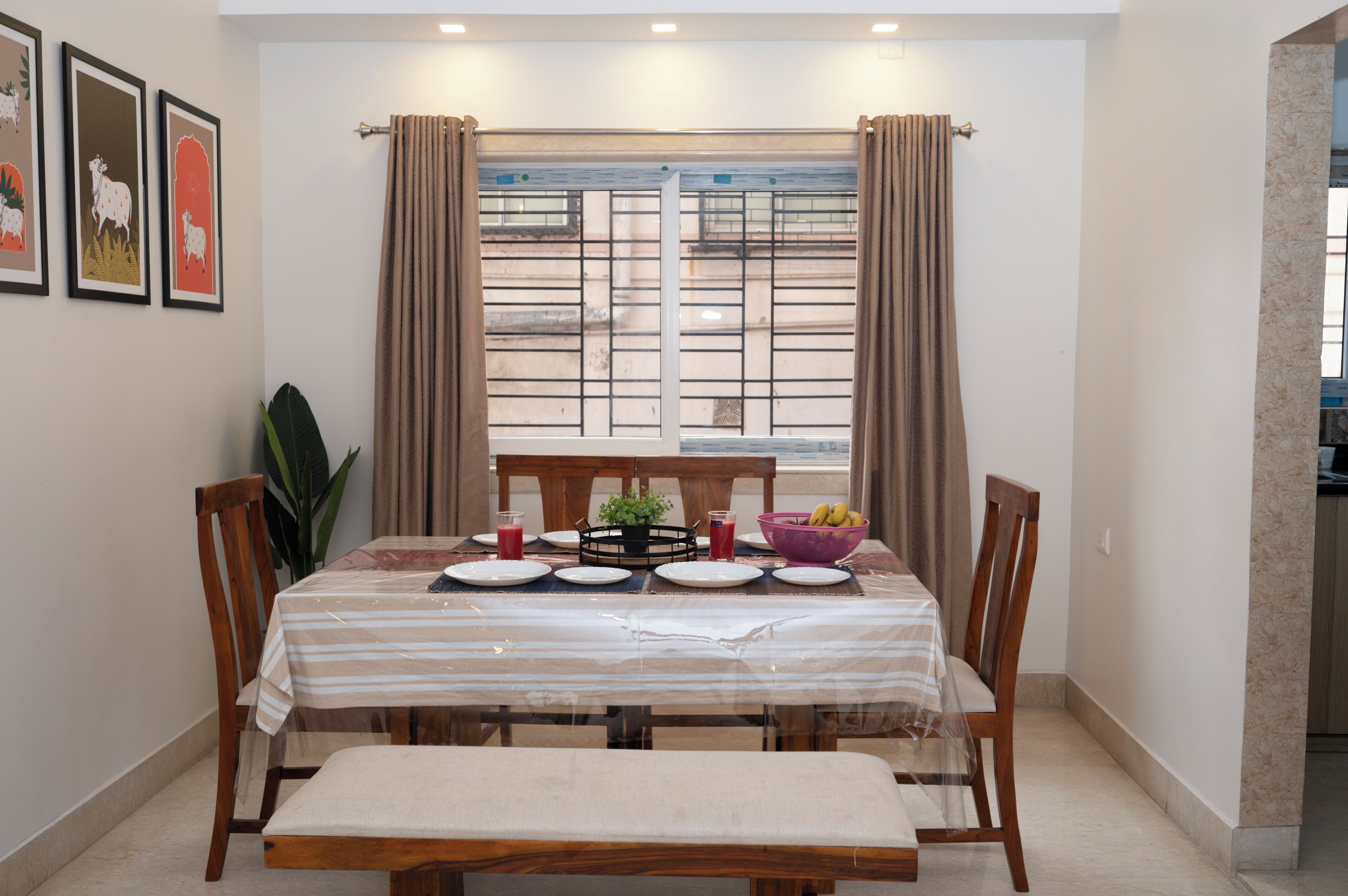 A dining area with a table set for four, a window with curtains, and framed artwork on the wall.