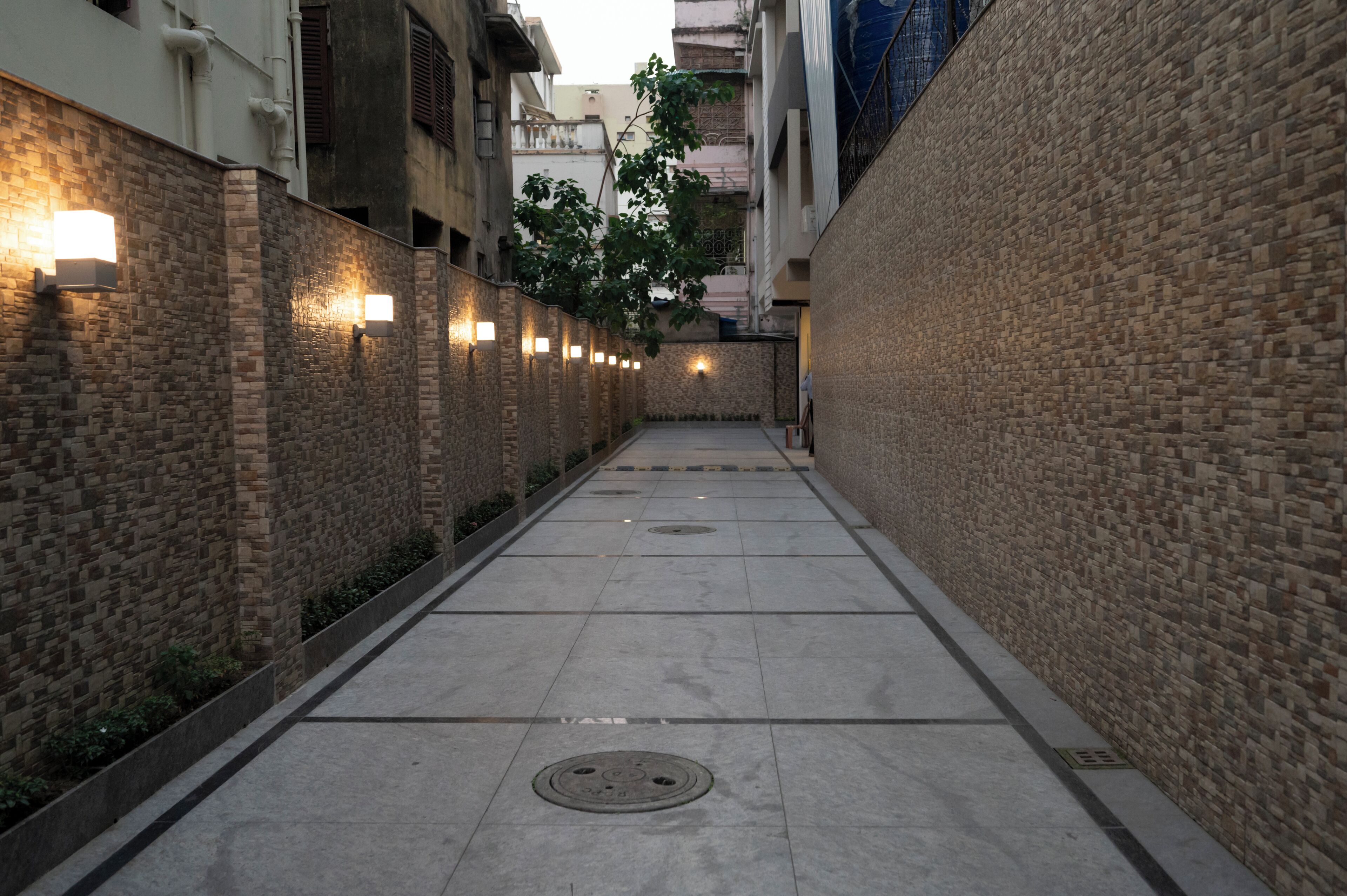 A narrow alleyway with brick walls and tiled flooring, featuring wall-mounted lights and a tree in the center.