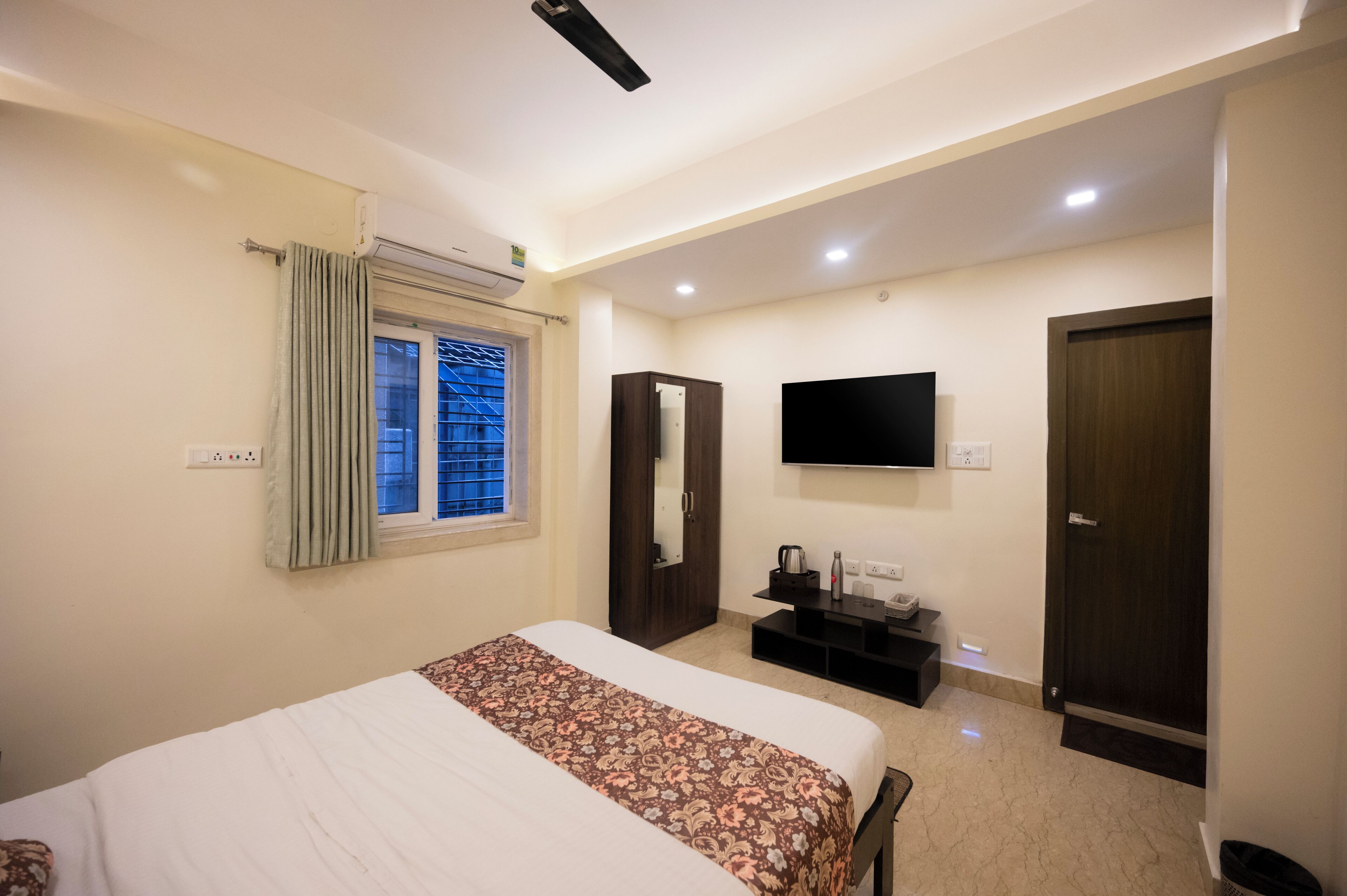 Habitación de hotel con cama, televisor montado en la pared, ventana con cortinas, armario y una mesita con hervidor y una botella.