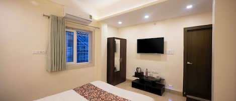 Ein Hotelzimmer mit einem Bett, einem an der Wand befestigten Fernseher, einem Fenster mit Vorhängen, einem Kleiderschrank und einem kleinen Tisch mit Wasserkocher und Flasche.