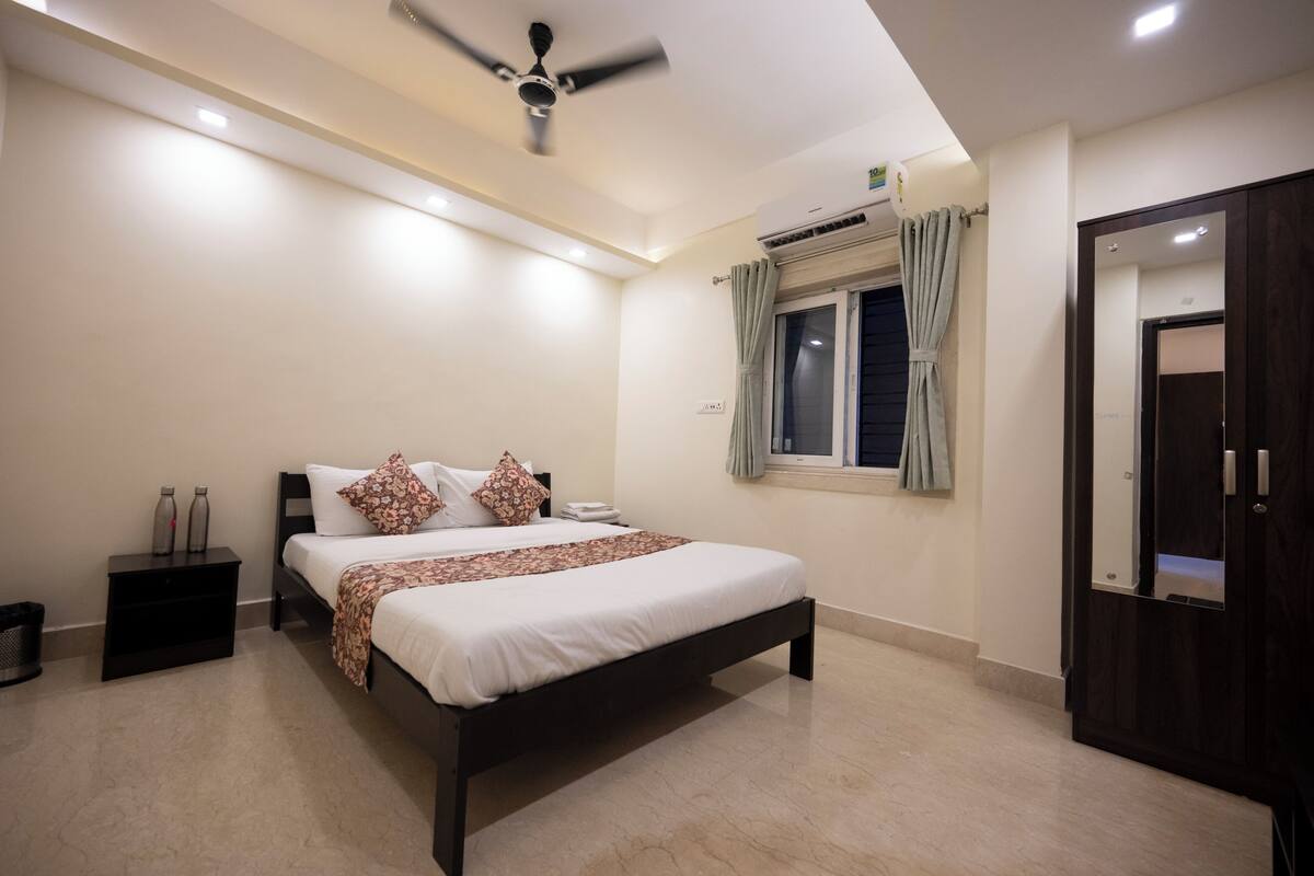 A bedroom with a bed, a ceiling fan, a window with curtains, a wardrobe, and a nightstand with bottles.