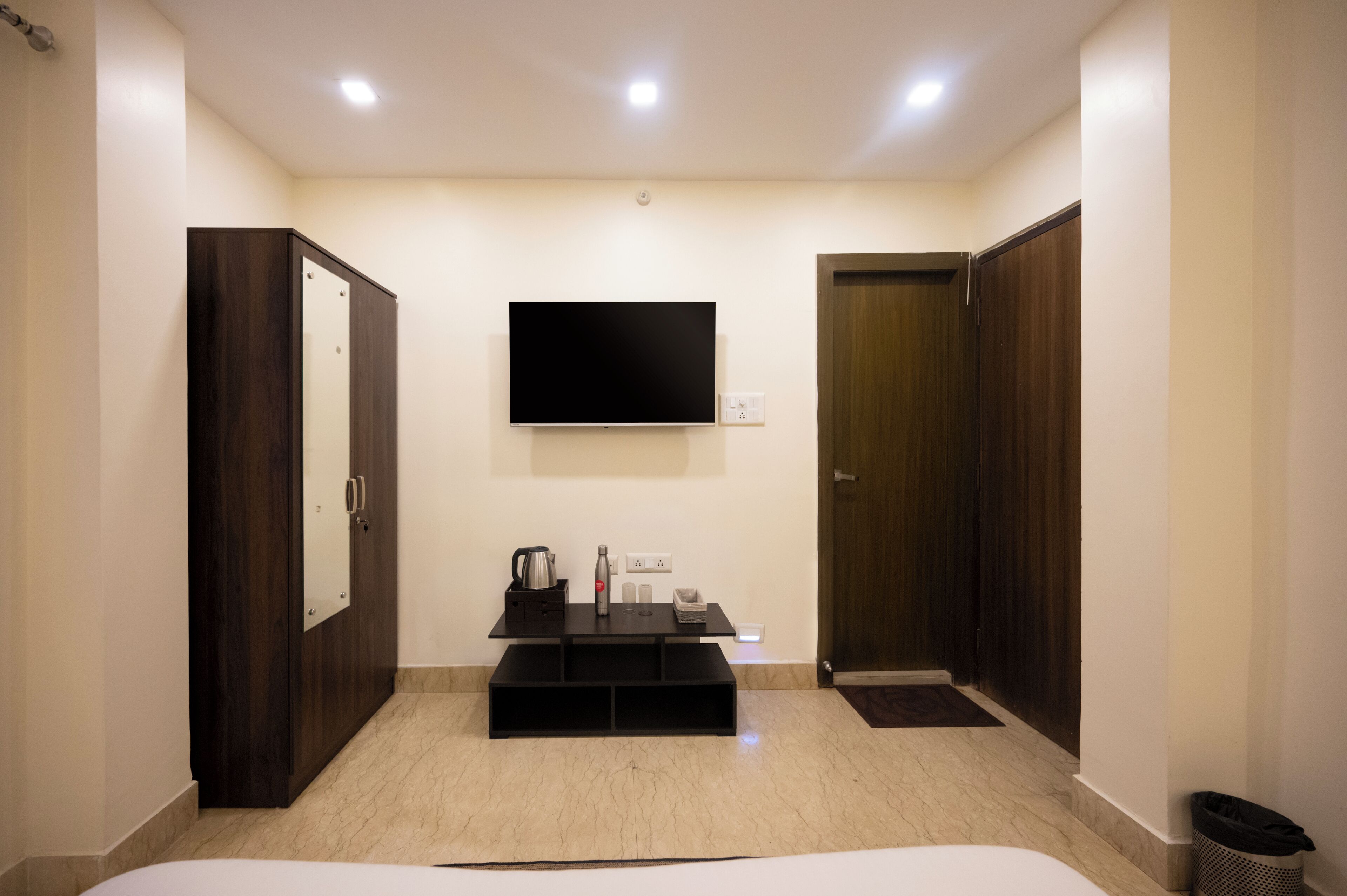 A modern hotel room with a flat-screen TV, a dark wooden wardrobe, a small table with a kettle and a bottle, and a trash bin.